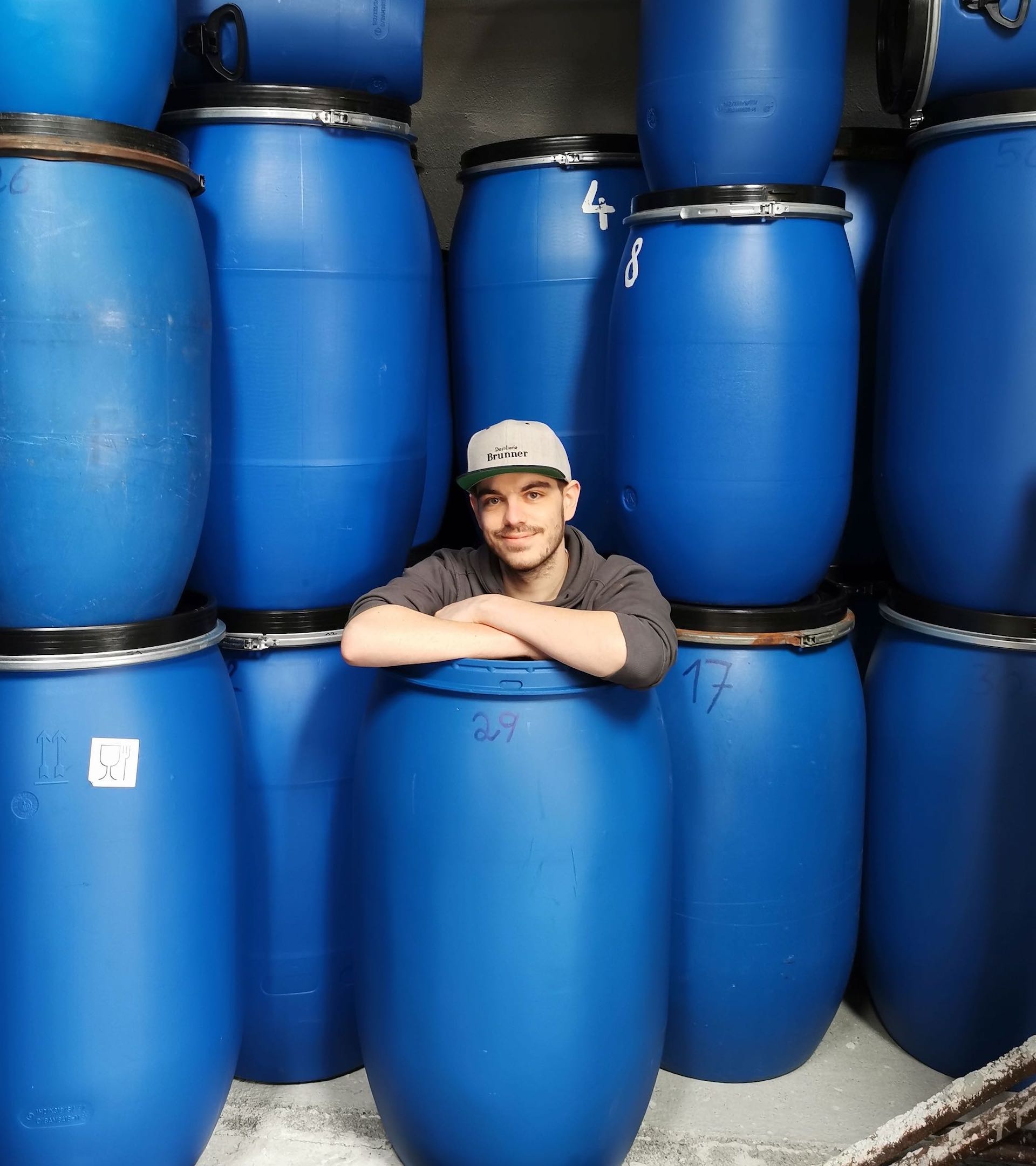 Daniel Brunner sitzend und lächelnd in blauen Maischefässern mit gebrandeter Destillerie Brunner Flexfit auf dem Kopf.